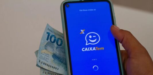 Foto mostra uma pessoa segurando um celular com o aplicativo do Caixa Tem e notas de dinheiro