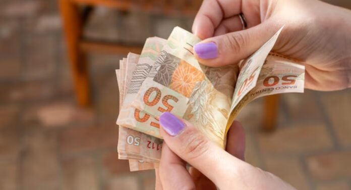 Foto mostra pessoa contando cédulas de dinheiro de auxílio do caixa tem