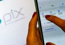 Imagem de uma mão segurando um celular com o app do banco aberto com um notebook ao fundo com a imagem do PIX