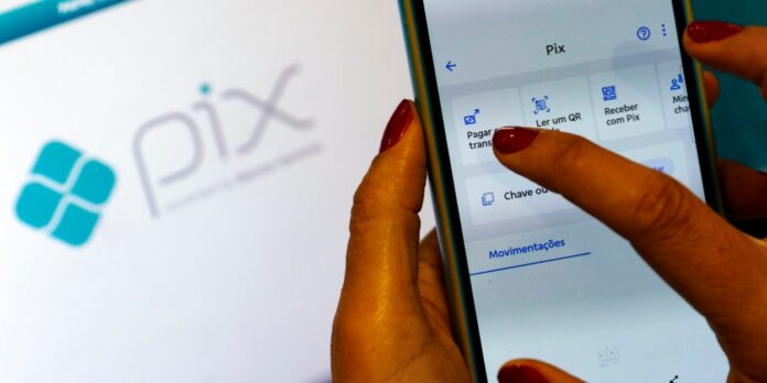 Imagem de uma mão segurando um celular com o app do banco aberto com um notebook ao fundo com a imagem do PIX