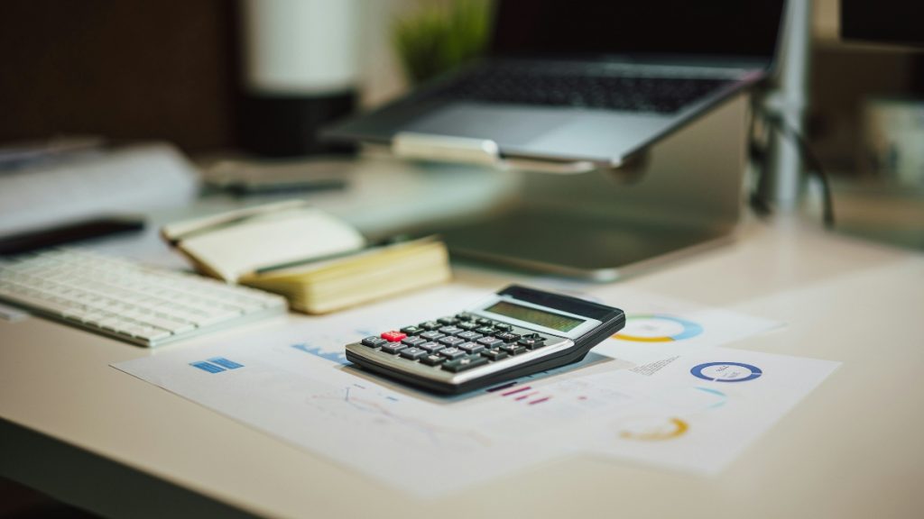  Calculadora sobre uma folha com gráficos financeiros em uma mesa de trabalho, ao lado de teclado, caderno e notebook, ilustrando o cálculo do custo efetivo total antes de assinar